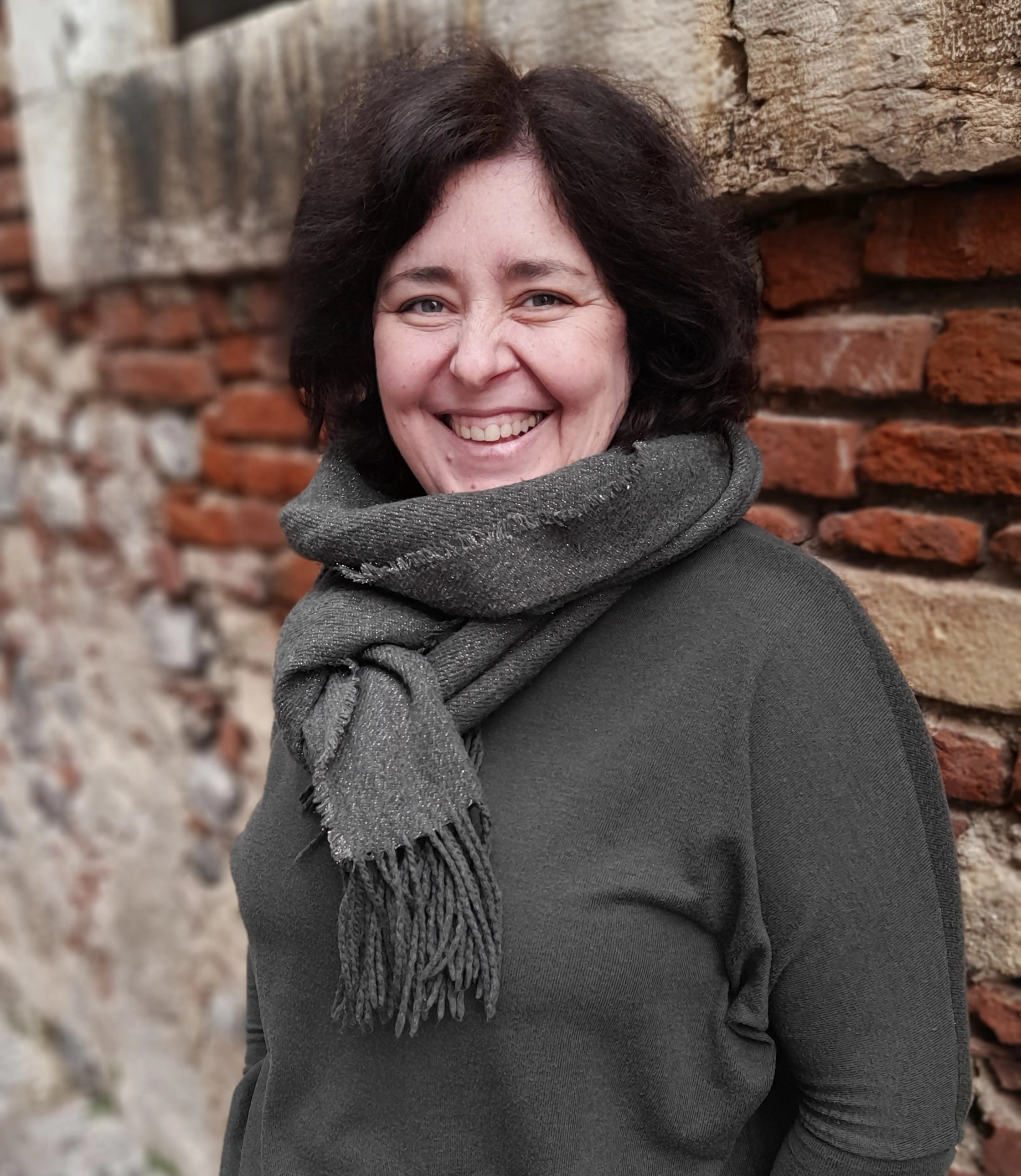 Photo of Tamara Tempera, on a brick wall background. Tamara is wearing a dark green sweater and matching scarf